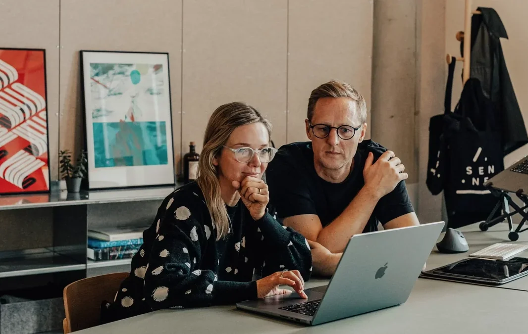 Two people working side by side at a laptop in the Sennep studio, below a wall of framed prints.