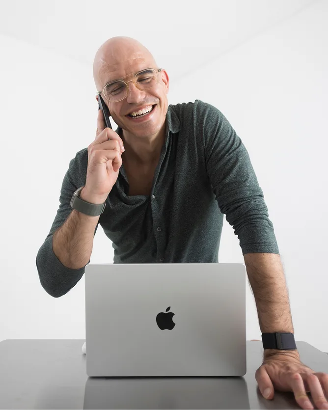 Fábio laughing on a phone call, leaning on a desk behind a silver laptop, in a dark green shirt against a white studio wall