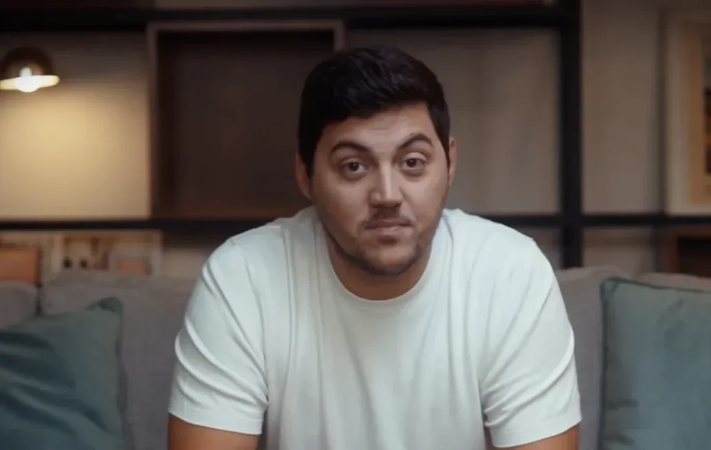 André Oliveira sitting on a sofa in a white t-shirt, looking slightly off-camera in a softly lit living room