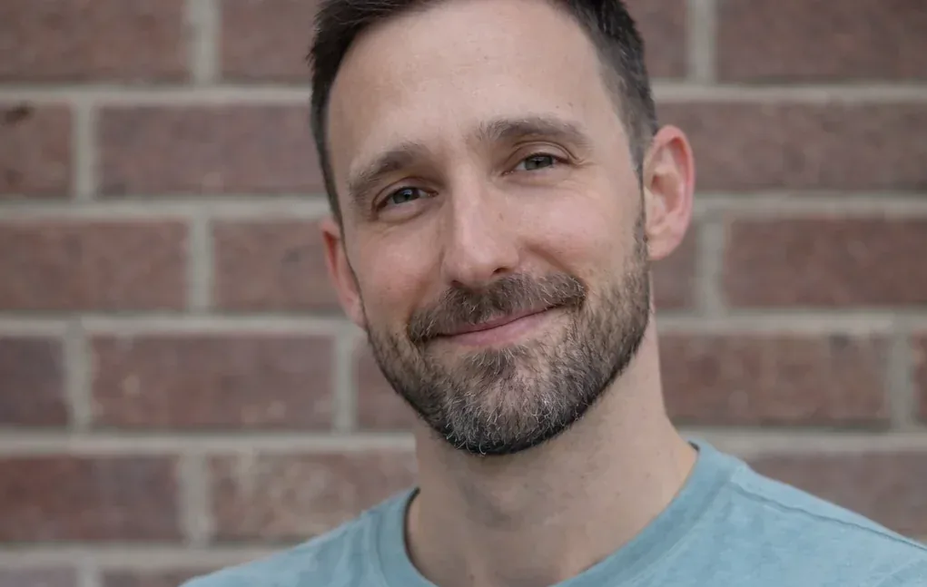 Marc Simon smiling outdoors in a light blue t-shirt against a brick wall